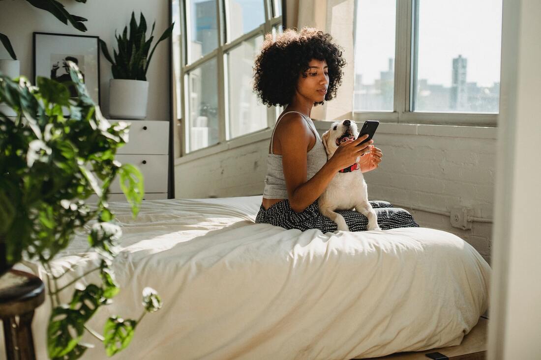 A woman with a smartphone and a dog sitting on a bed in the house.
