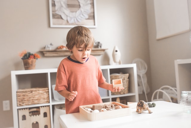 Boy wearing a red sweatshirt playing with blocks in a room with an open shelf full of toys behind him